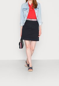 Giacca di jeans, polo rossa, gonna blu navy con cintura a strisce rosse e bianche, borsa nera con dettagli rosso-bianco-blu, sandali blu navy.