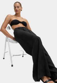 Czarny top bandeau z detalem w postaci węzła, w komplecie z czarnymi, wysokim stanem spodniami o szerokich nogawkach. Gładka tekstura materiału, noszony z minimalistycznymi czarnymi sandałami.