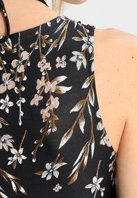 Kwiatowy top bez rękawów z czarnym tłem, pokryty białymi i brązowymi kwiatami, o fakturowanej powierzchni i czarnym pasku na szyi.