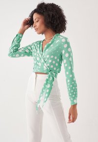 Grünes Wickeloberteil mit Punkten aus leichtem Stoff, langärmlig und mit einem Bindeband in der Taille; kombiniert mit hoch taillierten weißen Hosen.