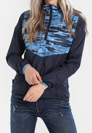 Personne portant une veste à motif camouflage bleu marine et bleu avec une fermeture éclair à moitié et des poignets élastiques, ajustant le poignet. Elle porte également un jean bleu foncé.