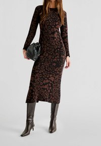 Langärmliges, tailliertes Kleid im schwarz-braunen Leopardenmuster mit Midi-Länge. Kombiniert mit einer schwarzen Tasche und kniehohen braunen Stiefeln.