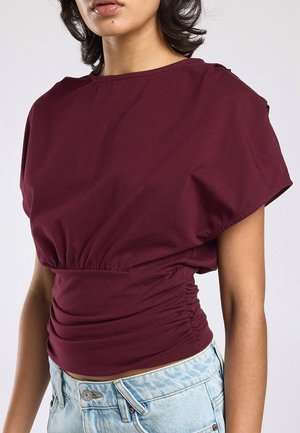 Vrouw draagt een bordeauxrood gerimpeld topje met korte mouwen en lichtblauwe hoog taille spijkerbroek, getoond van schouders tot heupen.