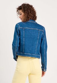 Chaqueta de mezclilla con lavado azul, cuello clásico y cierre de botones. La parte trasera presenta costuras visibles y se combina con pantalones amarillo claro.
