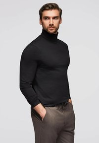Schwarzes Rollkragenoberteil, taillierte Passform, lange Ärmel. Kombiniert mit hellbraunen Hosen, Taschen sichtbar, glatte Textur, neutraler Hintergrund.