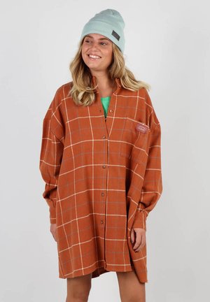 Chemise à carreaux orange surdimensionnée avec des manches longues, devant boutonné et une poche poitrine. Portée par-dessus un haut vert, associée à un bonnet bleu clair.