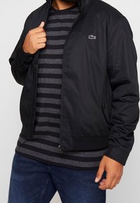 Man som bär en svart zip-up jacka över en grå och svart randig skjorta med blå jeans, håller i kragen på jackan med en hand.