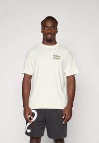 Lys beige t-shirt med korte ærmer og et lille grønt logo på brystet, parret med mørke shorts med en stor hvid grafik.