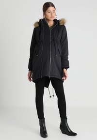 Svart polstret parka-jakke med hette trimmet med kunstpell, justerbar snor i livet, og glidelås foran, kombinert med svarte leggings og ankelboots.