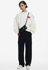 Felpa over-size color crema con grafica rossa, borsa a tracolla nera con borchie, pantaloni cargo neri e sneakers bianche con dettagli gialli.