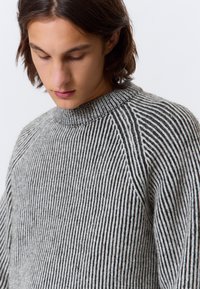 Jeune personne aux cheveux bruns mi-longs portant un pull en tricot gris et noir à rayures verticales, regardant vers le bas sur un fond blanc.