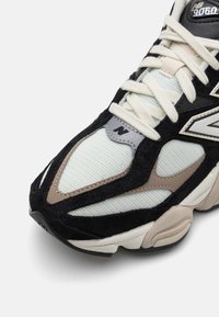 Gros plan d'une sneaker New Balance 9060 avec des panneaux en mesh et en suède noirs, blancs, gris et beige, et des lacets blancs.