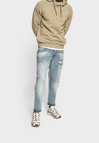 Hoodie beige con un design testurizzato, abbinato a jeans chiari strappati con un accento di catena metallica e sneakers bianche con dettagli multicolori.