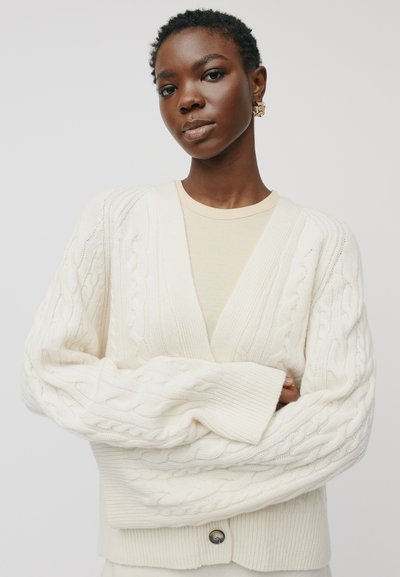 Jauna juoda moteris su trumpais natūraliais plaukais dėvi grietinėlės spalvos pintą megztinį virš šviesiai kreminės spalvos palaidinės, su sukryžiuotomis rankomis, auksinėmis auskarais, neutralus fonas.