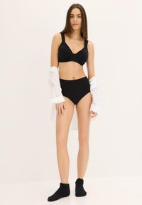 Reggiseno nero e pantaloni alti in vita realizzati in tessuto liscio, indossati con una camicia bianca. La modella indossa calzini neri pelosi alla caviglia.