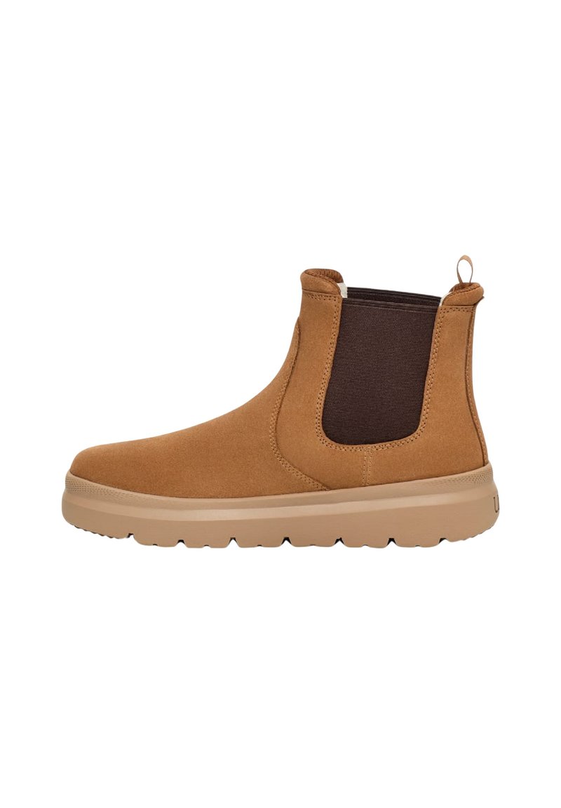 Botín de ante marrón a la altura del tobillo con paneles laterales elásticos, puntera redonda y suela de goma beige. Incluye una asa en el talón para facilitar el uso.
