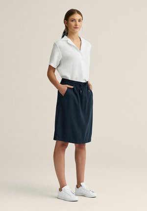 Kvinde iført en hvid kortærmet bluse, mørkeblå nederdel til knæet med lommer, hvide sneakers og et mønstret hårtørklæde, stående med hænderne i lommerne.