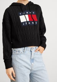 Person wearing a black cable-knit hoodie with Tommy Jeans logo and light blue high-waisted jeans, hand in pocket, against white background.