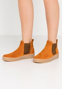 Nogawki ubrane w brązowe zamszowe botki za kostkę z elastycznymi panelami bocznymi i beżowymi gumowymi podeszwami na gładkiej białej podłodze.