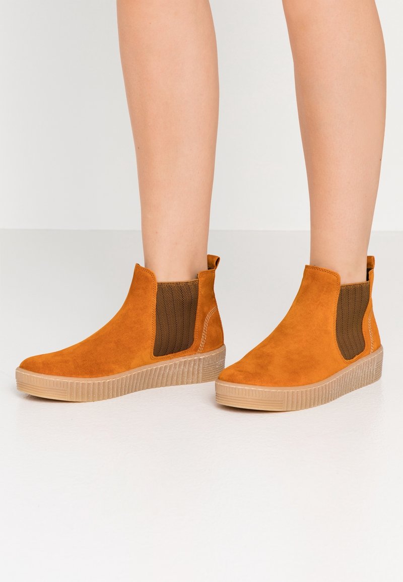 Nogawki ubrane w brązowe zamszowe botki za kostkę z elastycznymi panelami bocznymi i beżowymi gumowymi podeszwami na gładkiej białej podłodze.