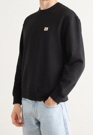 Mężczyzna ubrany w czarną bluzę Calvin Klein i jasnoniebieskie dżinsy, z jedną ręką w kieszeni, na tle jednolitego jasnego tła.