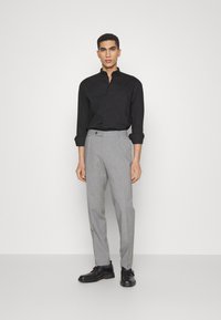 Camicia nera con colletto a bottoni, pantaloni grigi su misura e scarpe nere. I pantaloni presentano un solo bottone e tasche laterali.