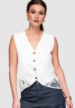 Frau trägt eine ärmellose weiße Weste mit goldenen Knöpfen, Spitzen-Details, dunkle Jeans, goldene Ohrringe und ein goldenes Armband vor neutralem Hintergrund.