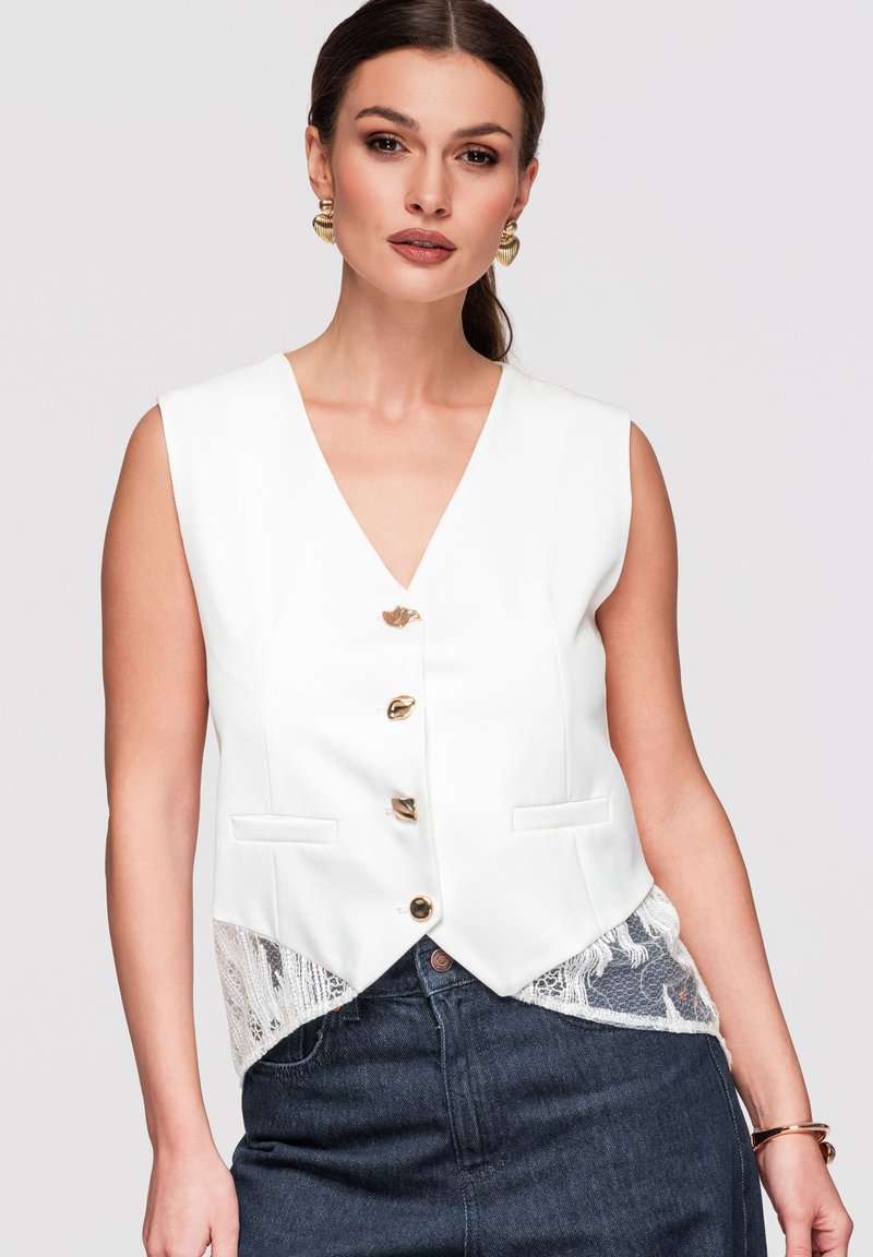 Femme portant un gilet blanc sans manches avec des boutons dorés, des détails en dentelle, un jean foncé, des boucles d’oreilles dorées et un bracelet doré sur un fond uni.