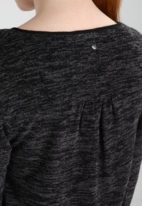 Svart strukturerad tröja med ett subtilt mönster, med en plisségång på baksidan och en dekorativ metallknapp nära halsen.