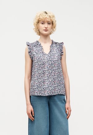 Νεαρός με σγουρά ξανθά μαλλιά, φορώντας αμάνικο φλοράλ μπλουζάκι και μπλε τζιν παντελόνι με φαρδιά πόδια, στέκεται μπροστά σε απλό φόντο.