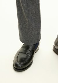 Svarta läderloafers med en slät yta, som har en spetsig tå och remdetalj. Staplade med grå skräddarsydda byxor.