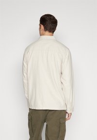 Licht beige overhemdjasje met een kraag, lange mouwen en knoopmanchetten. Gestoft stof met een relaxte pasvorm en een achterpand detail.
