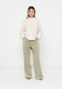 Chunky cremefarbener Strickpullover mit Fransenverzierung, kombiniert mit weit geschnittenen hellgrünen Jeans. Sanfte Textur, entspannter Schnitt, schlichtes Design.