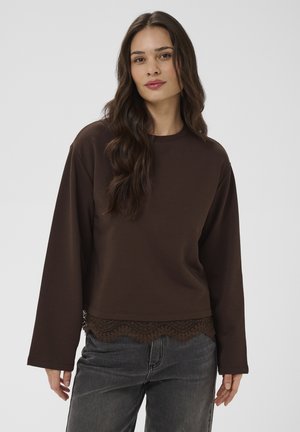 Femme aux longs cheveux bruns portant un haut ample à manches longues marron foncé avec bordure en dentelle et un jean gris, debout devant un fond uni.