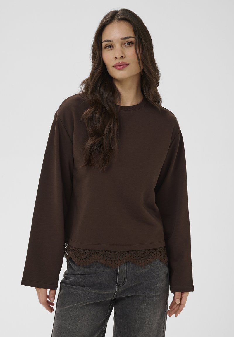 Femme aux longs cheveux bruns portant un haut ample à manches longues marron foncé avec bordure en dentelle et un jean gris, debout devant un fond uni.