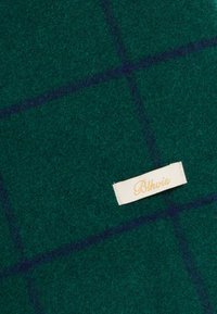 Grønt ullteppe med et mørkeblått rutemønster. En kremfarget etikett med gullskrift som sier "Blkvis." Myk tekstur med en varm utseende.