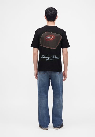Άνδρας που φοράει μαύρο T-shirt με πολύχρωμη σκηνή κινηματογράφου και το κείμενο "Filling Pieces Art of Cinema" στην πλάτη, σε συνδυασμό με μπλε τζιν και αθλητικά παπούτσια.
