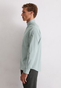 Camicia button-up verde chiaro con colletto piccolo, tasca sul petto e maniche lunghe. La texture appare liscia con una vestibilità rilassata.