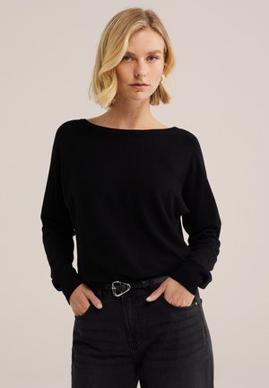 Blonde Frau trägt ein schwarzes Langarmoberteil und schwarze Jeans mit hoher Taille und silberner Gürtelschnalle, Hände in den Taschen, neutraler Hintergrund.