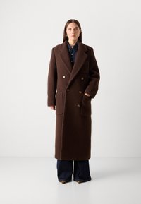 Cappotto doppio petto in lana marrone caratterizzato da una silhouette lunga, ampi rever e tasche frontali, abbinato a una camicia in denim blu navy e jeans svasati.