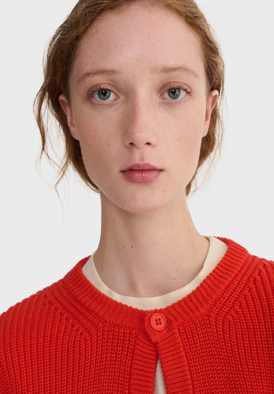 Jeune femme à la peau claire, aux yeux bleu clair et aux cheveux châtain clair portant un pull en laine rouge vif par-dessus une chemise crème, regardant droit devant.