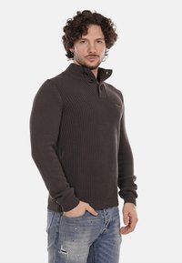 Maglione lavorato a maglia marrone scuro con colletto alto, pattina con due bottoni e polsini a costine, abbinato a jeans blu sbiaditi.