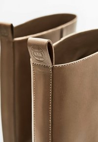 Close-up van tan leren laarzen met trekbanden en zichtbare stiksels tegen een effen witte achtergrond.