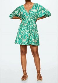 Femme portant une robe courte verte à fleurs avec des manches bouffantes et des sandales marron à lacets, debout les mains sur les hanches.