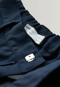 Tissu bleu marine avec une surface texturée, comportant une étiquette indiquant "NOM" et une fermeture à fermoir argenté. Le design comprend des bords froncés.