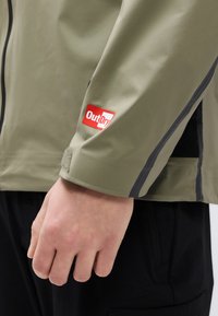 Κοντινό πλάνο του χεριού ενός ατόμου δίπλα σε μανίκι πράσινου λαδί μπουφάν με κόκκινο και άσπρο λογότυπο OutDry και ορατό μαύρο παντελόνι.