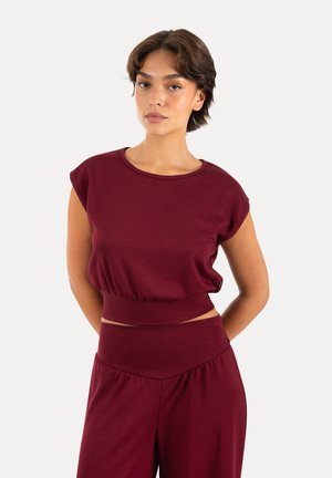 Mujer con cabello corto y oscuro que lleva una blusa sin mangas color granate y pantalones de talle alto a juego, de pie frente a un fondo claro y sencillo.