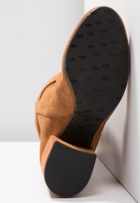 Bottine en daim couleur camel avec un bout arrondi, un petit talon bloc et une semelle extérieure en caoutchouc noir texturé, ornée de logos embossés et de détails de taille.