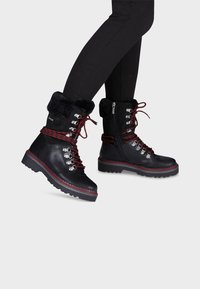 Botas de cuero negras hasta el tobillo con cordones rojos y negros, cuello de pelo sintético, herrajes plateados y suela resistente. Cierre de cremallera en un lado para facilitar su uso.