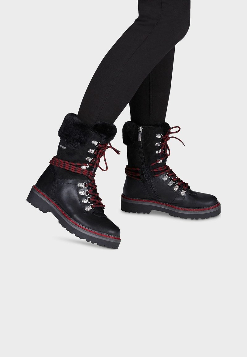 Botas de cuero negras hasta el tobillo con cordones rojos y negros, cuello de pelo sintético, herrajes plateados y suela resistente. Cierre de cremallera en un lado para facilitar su uso.